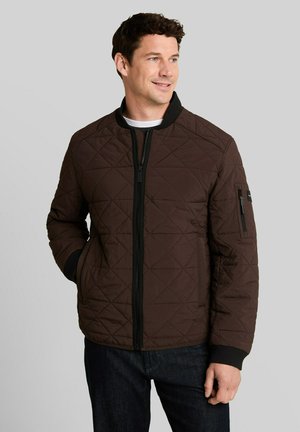 Man wearing a dark brown quilted jacket with black zipper and cuffs, standing with hands in pockets, looking slightly to the side and smiling.