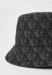 Cappello a secchiello nero con un motivo testurizzato di lettere intrecciate. Realizzato in tessuto con una leggera lucentezza, presenta una visiera larga e flessibile.