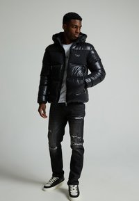 Schwarze Daunenjacke mit glänzender Oberfläche, Reißverschluss vorne und Kapuze. Trägt man über einem grauen Shirt und kombiniert mit verwaschenen schwarzen Jeans und schwarzen Sneakers.