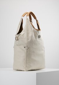Beige tote bag med struktureret trekantet mønster, brune polstrede håndtag, sidelommer og et rundt metallogo emblemer på fronten.