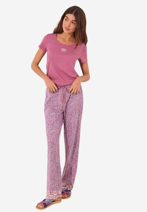 Roze T-shirt met een klein wit logo, gecombineerd met ruime, gestreepte broeken in roze en wit, aangevuld met blauwe en paarse slippers.