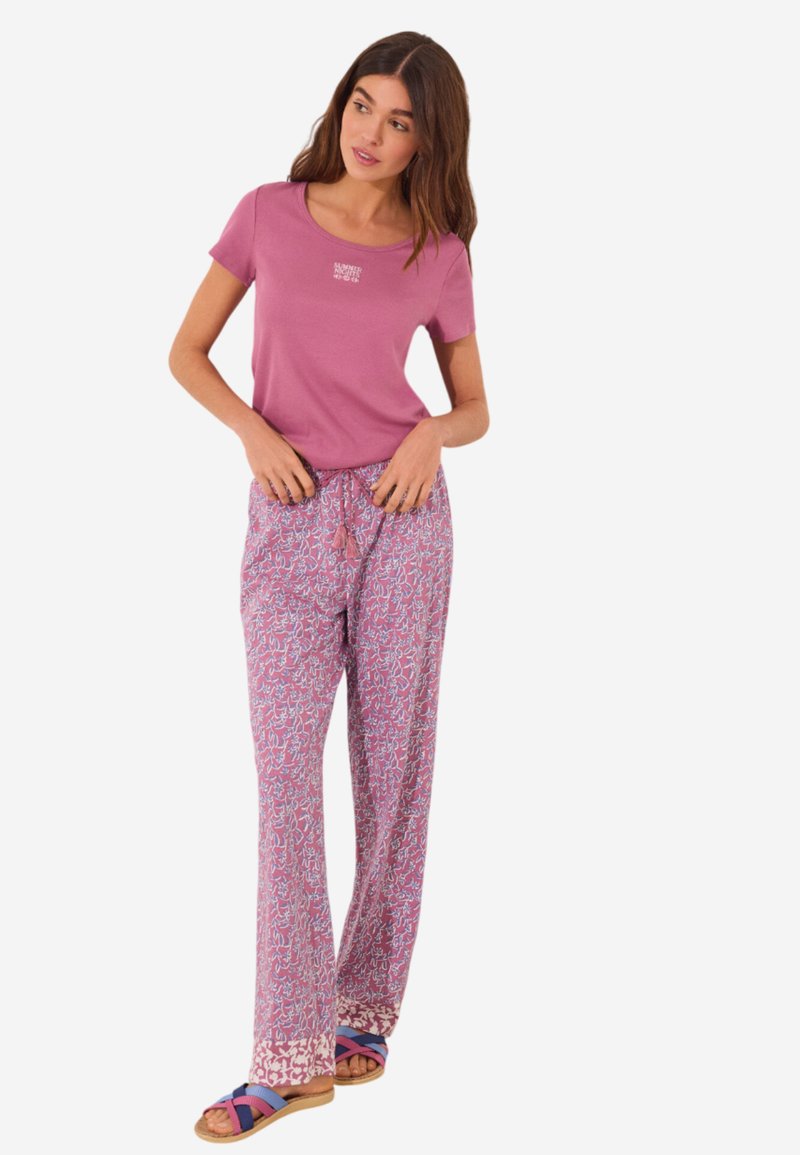 Rosa T-Shirt mit einem kleinen weißen Logo, kombiniert mit gemusterten, weiten Hosen in Rosa und Weiß, akzentuiert mit blauen und lila Schuhen mit offenem Zehen.
