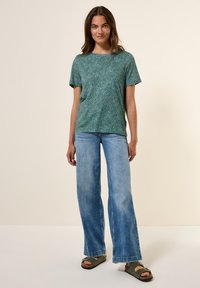 Chemise verte à manches courtes avec un motif en vagues, associée à un jean évasé bleu clair et des sandales ouvertes. Coupe décontractée et style casual.