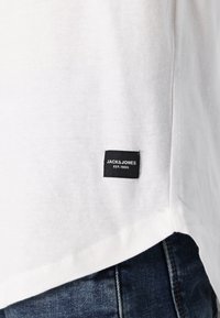 T-shirt en coton blanc avec une étiquette noire affichant "JACK & JONES EST. 1990" au bas, présentant un bord inférieur incurvé et des coutures décoratives.