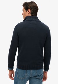 Marineblauer Strickpullover mit hohem, geripptem Kragen, eng anliegenden Ärmeln und geripptem Saum. Kombiniert mit hellblauen Jeans und zeigt einen lässigen Stil.