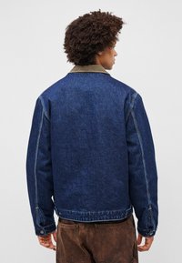 Jachetă din denim de culoare albastru închis, cu guler din corduroy bej. Prezintă o croială lejeră, buzunare laterale și detalii de cusătură vizibile.