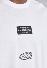 Fehér pamut póló, fekete hímzett folttal, amelyen az "Armani Exchange 1991" felirat és az "AX" logó nyomtatva van a hemnél. Sima textúrájú.