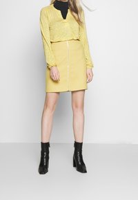 Gelbe gemusterte Bluse mit langen Ärmeln, kombiniert mit einem gelben Bleistiftrock mit einem Reißverschluss an der Vorderseite und schwarzen Knöchelstiefeln.