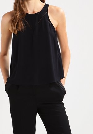 Femme portant un haut noir sans manches à col halter et un pantalon noir, debout avec les mains dans les poches devant un fond uni.