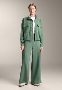 Zelena jakna z dvema sprednjima žepoma, zadrgo in stojnim ovratnikom, v kombinaciji s širokimi zelenimi hlačami. Gladek material, sproščen kroj in sproščen stil.