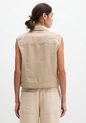 Femme aux cheveux foncés attachés en arrière portant un gilet beige sans manches et un pantalon beige assorti, vue de dos sur un fond blanc.