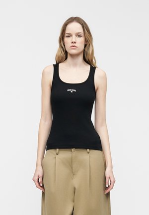 Joven con cabello rubio largo que lleva una camiseta sin mangas negra con el logo "GUESS JEANS" y pantalones beige de cintura alta, posando contra un fondo blanco.