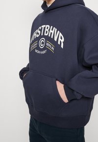 Sudadera azul marino de algodón suave, con un bolsillo delantero, puños acanalados y un diseño de texto en blanco y amarillo en el pecho.