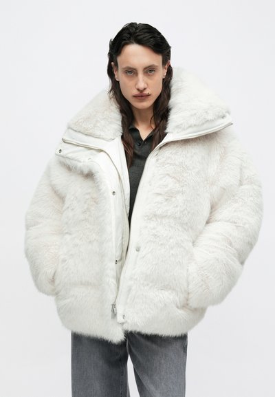 Veste matelassée blanche avec texture en fausse fourrure, grand col, fermeture éclair et boutons-pression. Portée sur une chemise sombre, associée à un jean gris.