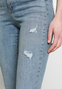 Světle modré džíny z rifloviny s přiléhavým střihem, které mají dvě odřená místa na horní části stehna a hladkou texturu.