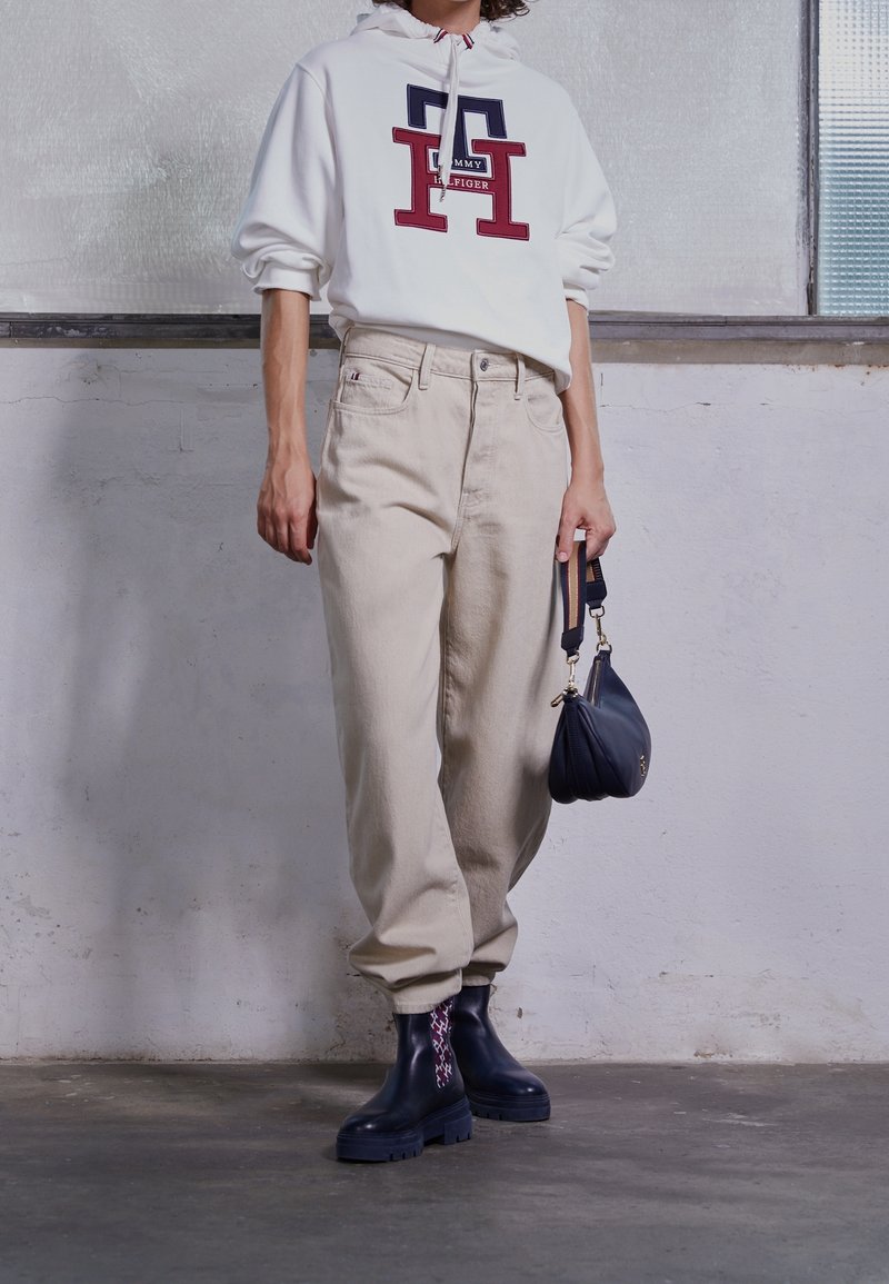 Personne portant un sweat à capuche blanc Tommy Hilfiger, un pantalon beige, des bottes noires avec des chaussettes à motifs, et tenant un petit sac noir.