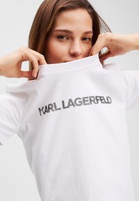 Balta medvilninė marškinėliai su dideliu sidabriniu "KARL LAGERFELD" logotipu krūtinės lygyje. Trumpi rankovės ir apykaklės dizainas. Lygi tekstūra.