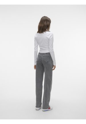 Femme debout de dos, portant un haut blanc à manches longues, un jean gris taille haute et des baskets blanches sur un fond uni.
