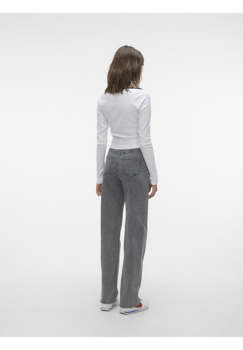 Femme debout de dos, portant un haut blanc à manches longues, un jean gris taille haute et des baskets blanches sur un fond uni.