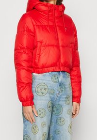 Giubbotto imbottito rosso con cappuccio, dotato di chiusura con zip, polsini elasticizzati e design corto. Indossato con pantaloni di jeans azzurri con stampe di faccine sorridenti.