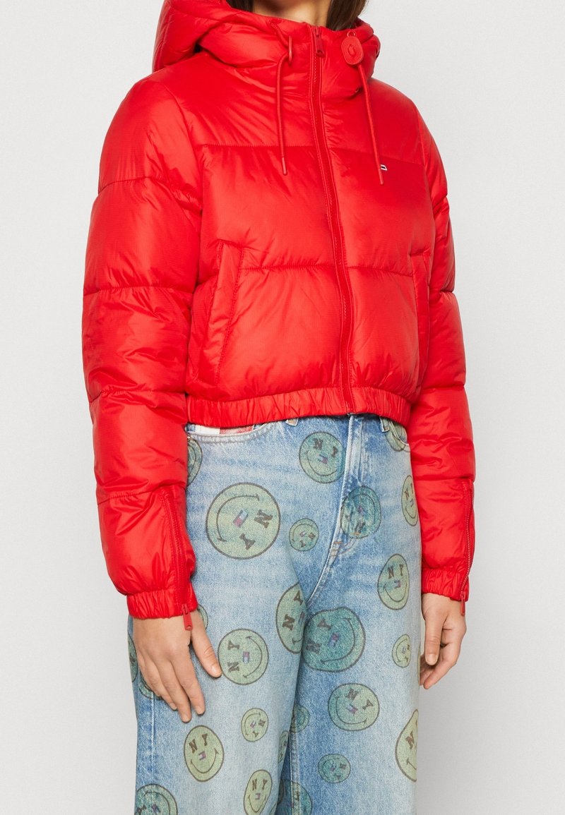 Giubbotto imbottito rosso con cappuccio, dotato di chiusura con zip, polsini elasticizzati e design corto. Indossato con pantaloni di jeans azzurri con stampe di faccine sorridenti.