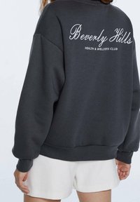 Sudadera gris de corte holgado, con texto bordado en blanco: "Beverly Hills CLUB DE SALUD Y BIENESTAR" en la parte posterior. Tejido de mezcla de algodón.