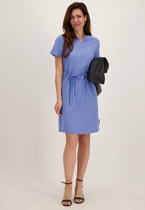 Vrouw in een knielange, korte mouwen blauwe jurk met een tailleceintuur, houdt een zwarte jas vast en draagt zwarte enkellaarzen met bandje.