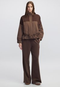 Bruine, twee-kleurige jas met een hoge kraag, een ingezette taille en elastische manchetten. Gecombineerd met losse, bruine broek en casual schoeisel.