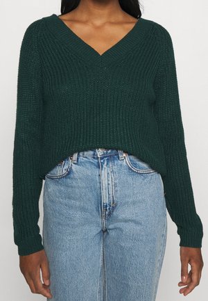 Ciemnozielony prążkowany sweter z dekoltem w serek, zestawiony z jasnoniebieskimi jeansami z wysokim stanem, widoczny od połowy klatki piersiowej do bioder.