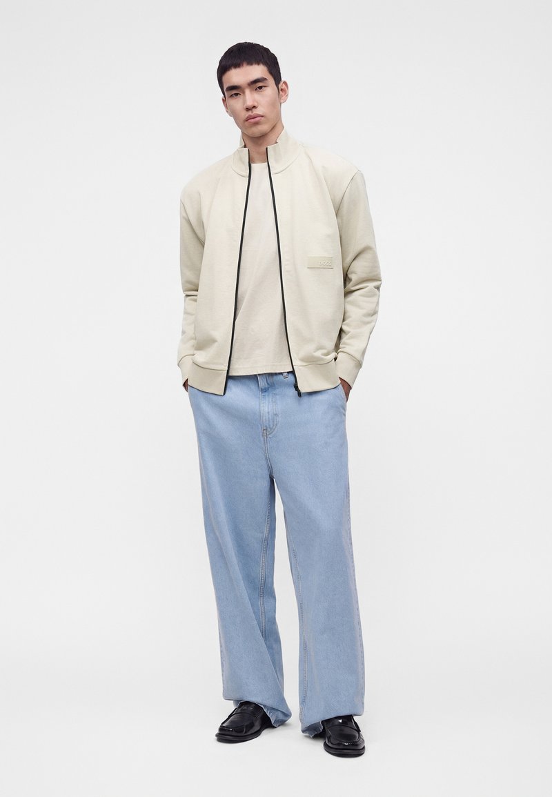 Junger Mann trägt eine beige Jacke mit Reißverschluss, hellblaue Jeans mit weitem Bein und schwarze Lederschuhe, steht mit den Händen in den Taschen.