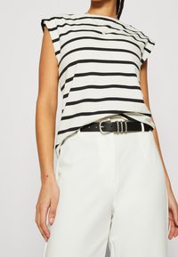 Camisa a rayas con rayas alternas en blanco y negro, escote redondo, mangas cortas, combinada con pantalones blancos y un cinturón negro.