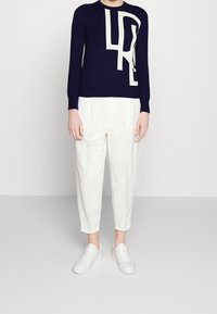 Marinblå stickad tröja med ett kontrasterande vitt abstrakt mönster, ihop med vita veckade byxor och vita sneakers.