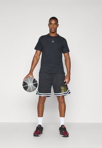 Camiseta atlética negra de manga corta con logo, pantalones cortos de baloncesto grises con el número amarillo "24" y zapatillas de baloncesto negro/rojas, sosteniendo un balón.
