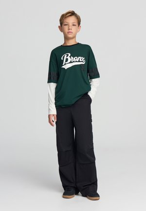T-shirt verde con grafica "Bronx" in bianco, sovrapposta a una maglietta bianca a maniche lunghe; abbinata a pantaloni cargo neri e scarpe nere.