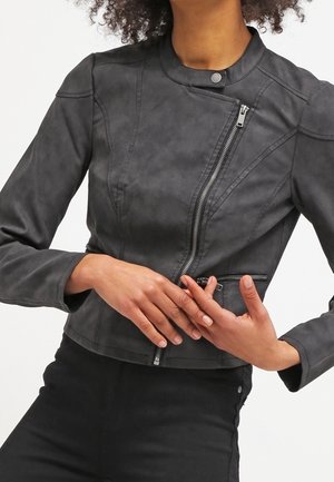 Frau trägt eine enganliegende schwarze Bikerjacke aus Kunstleder mit asymmetrischem silbernem Reißverschluss und Kragen mit Druckknopf, die Hände vor der Taille verschränkt.