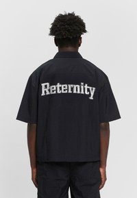 Schwarzes Kurzarmhemd mit strukturiertem Finish, auf dem Rücken mit großen weißen "Reternity"-Buchstaben bestickt.