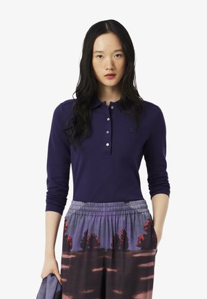 Polo a maniche lunghe in cotone viola scuro, con una chiusura a bottoni e un piccolo logo, abbinato a pantaloni colorati e con motivo.