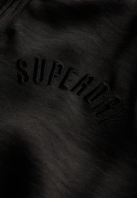 Zwarte stof met een gladde textuur heeft geborduurde tekst "SUPERDRY" in een bijpassende donkere kleur. Eenvoudig, minimalistisch ontwerp.