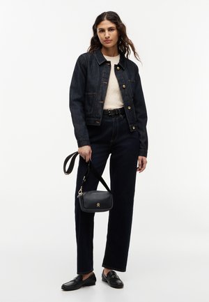 Frau trägt dunkelblauen Jeansjacke und Hose, cremefarbenen Pullover, schwarzen Gürtel, schwarze Loafers, hält eine schwarze Handtasche mit goldener Schnalle.