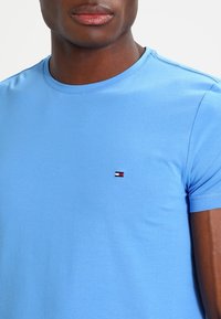 Světle modré tričko s krátkým rukávem vyrobené z měkké bavlny, s malým logem na hrudi s červenými, bílými a modrými doplňky.