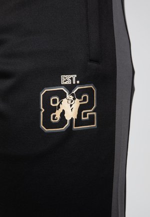 Zwarte joggingbroek met grijze zijpanelen, voorzien van een gouden grafisch ontwerp van "EST. 82" en een dierenillustratie op de linkerdij.