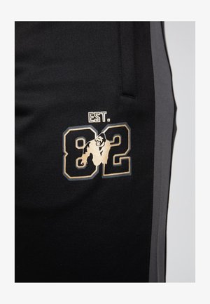 Zwarte joggingbroek met grijze zijpanelen, voorzien van een gouden grafisch ontwerp van "EST. 82" en een dierenillustratie op de linkerdij.