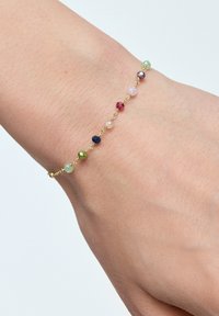 Złoty łańcuszek bransoletka z różnorodnymi kolorowymi, fasetowanymi koralikami w odcieniach zieleni, różu, czerwieni, niebieskiego i pomarańczowego, na tle skóry.