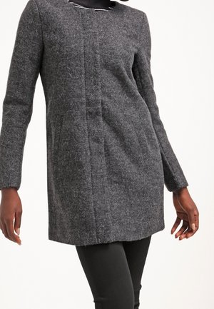 Femme portant un manteau en laine gris foncé ajusté avec des fermetures dissimulées à l'avant et des poches latérales, associé à un pantalon noir et une écharpe noire.