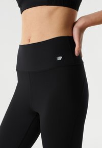 Zwarte sportleggings met een hoge tailleband. Gemaakt van gladde stof, met een klein wit "TWIST UP" logo op de heup.