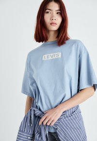 Lyseblå bomuld t-shirt med et hvidt Levi's logo på brystet, parret med en stribet skjorte med knapper, der er bundet om taljen.
