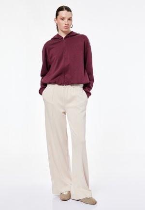 Kvinde iført en bordeaux rød lynlåshættetrøje, beige bukser med vide ben og beige sko, stående med hænderne i lommerne mod en hvid baggrund.