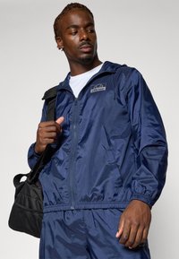 Marineblauwe windbreakerjas met elastische manchetten en zoom, voorzien van een logopatch. Accessoires omvatten een zwarte schoudertas. Glad, glanzend materiaal.