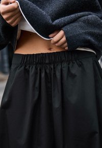 Fekete, magasderekú szoknya rugalmas derékpánttal; sima textúrájú, enyhe fényű. A modellen sötét kötött pulóver a derék fölött.