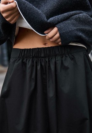 Fustă neagră, cu talie înaltă, elastică în talie; textură netedă, cu o ușoară strălucire. Modelul poartă un pulover tricotat închis la culoare deasupra taliei.